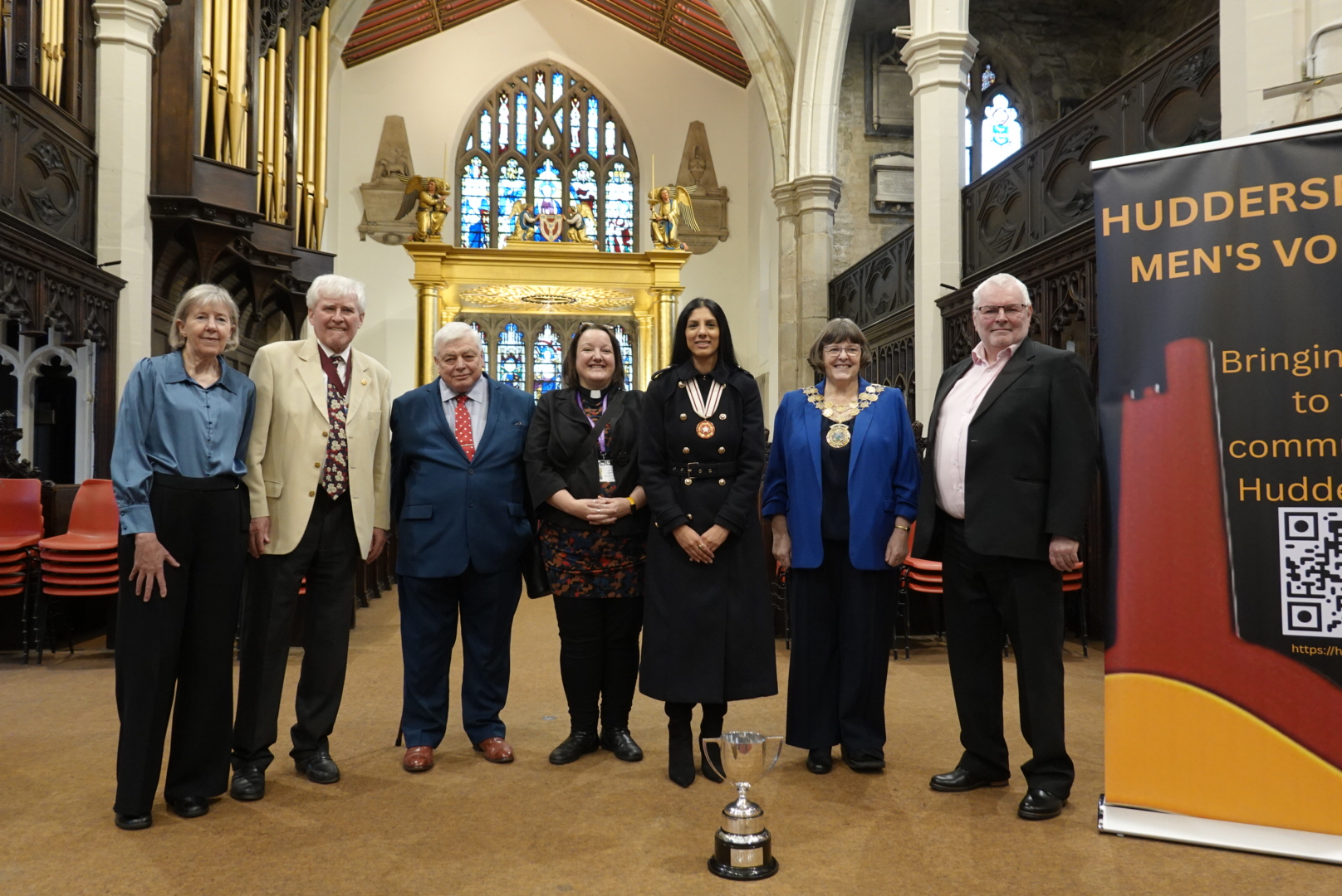 James and Jane standing with a number of guests inside the Church. To the right is the Choir's banner stand and the trophy is positioned on the floor.