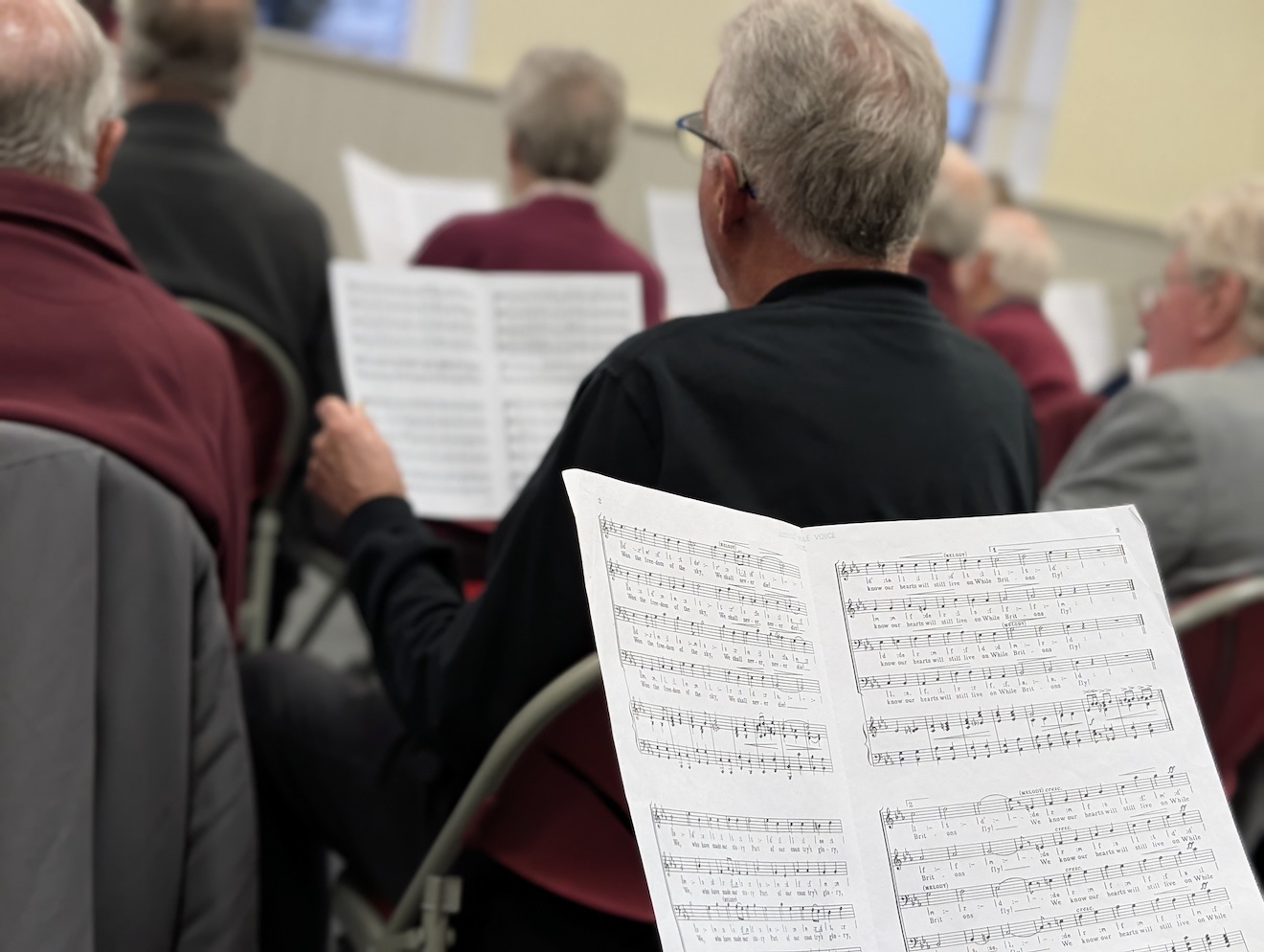 17:Choir rehearsal. Photo is a close up of choir members reading their music sheets.