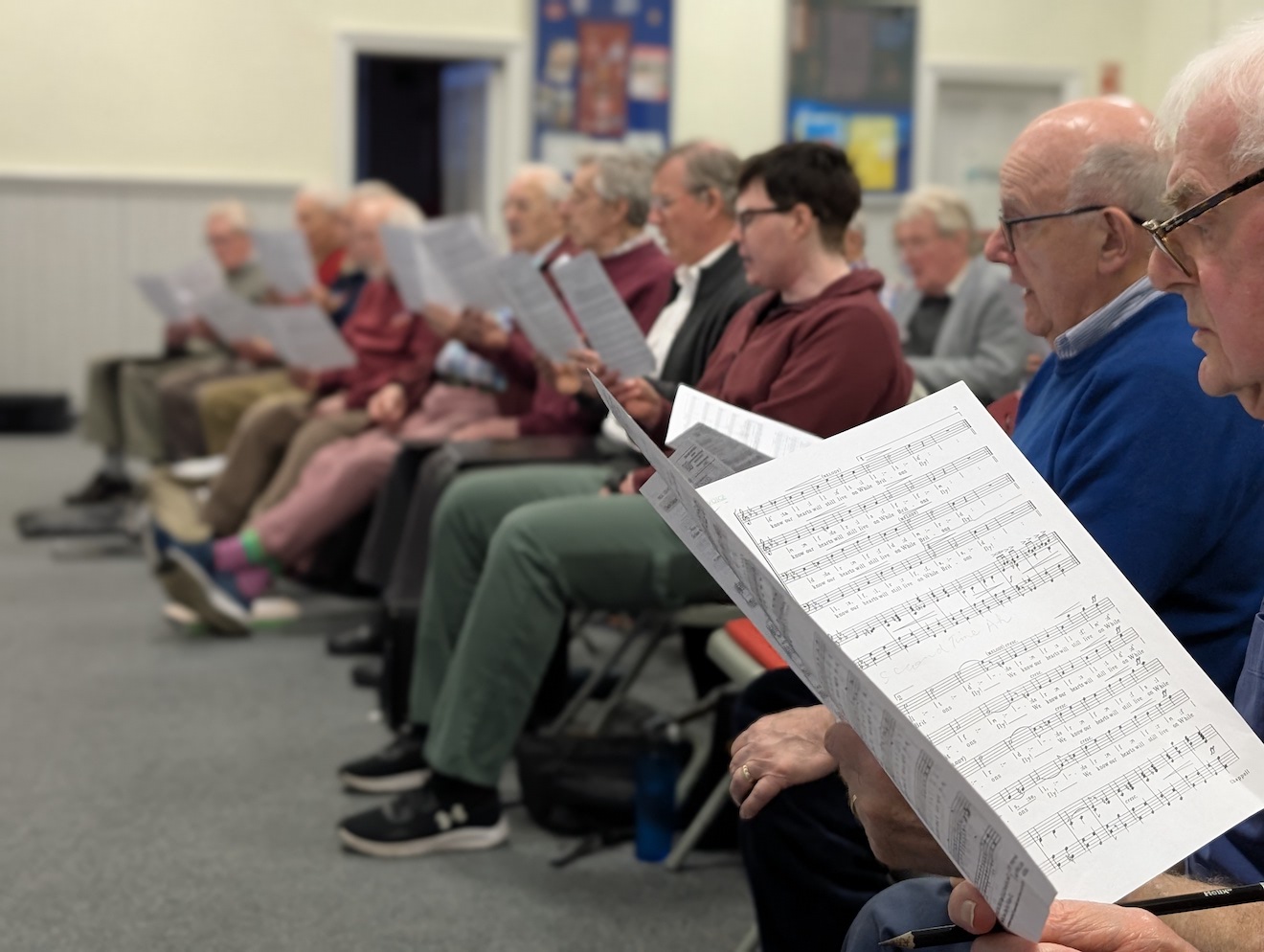 18:Choir rehearsal. Photo is a close up from the side of the room of choir members reading their music sheets.